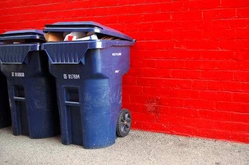 Two large dark blue plastic wheelie bins with black lids are positioned on a concrete surface in front of a bright red brick wall. The bins, which are used for rubbish collection, are slightly weathered and have visible white labels or markings on their sides. The bin to the right has some contents visible on top, including paper and possibly plastic waste, peeking from under the lid. The wheels of the bins are prominently visible, with the right bin’s wheel slightly turned outward. The scene appears to be outdoors, likely on a residential driveway or pavement area in Shepherds Bush, with natural lighting casting soft shadows. The vivid red wall provides a contrasting backdrop that emphasizes the bins’ utilitarian appearance, consistent with commercial waste collection and rubbish removal services by Commercial Waste Shepherds Bush.